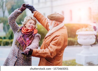 Happy Old Couple Dancing At Autumn Park. Senior Ma Flirting With Blonde Elderly Woman Outdoors In City Square In Cold Weather City Street. My Love Let's Dance Together. Lifestyle Invitation Concept.