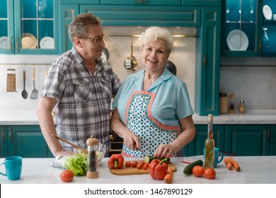 Happy Old Couple In Kitchen. Thankful Husband Hugs His Wife Cooking A Meal With Fresh Veggies.