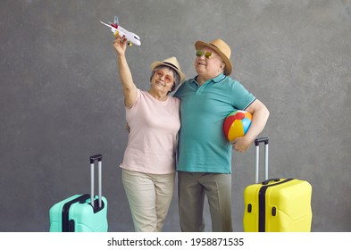 Happy old couple making dream come true. Smiling senior tourists in sunglasses and sun hats holding paper passenger airplane standing on grey studio background. Air flight and sea beach holiday