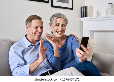 Happy Old Couple Waving Hands Making Online Video Call On Cell Phone. Mature Retired 50s Grandparents Enjoying Virtual Meeting Videocall Chat, Talking, Looking At Smartphone Sitting On Couch At Home.