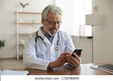 Happy Old Doctor Holding Smart Phone Texting Message Video Calling For Online Telemedicine Consultation In Medical Office. Smiling Senior Male Physician Using Mobile Healthcare App On Cell Technology.