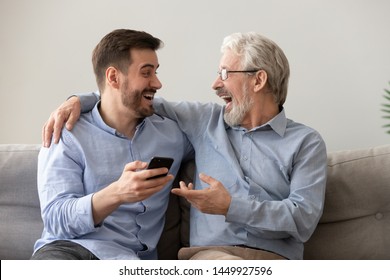 Happy Old Father And Son Having Fun At Home, Laughing, Using Phone Together, Mature Senior Dad And Millennial Man Discussing Funny News In Social Network Or Watching Video Online, Sitting On Couch