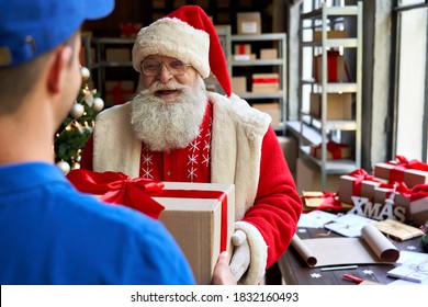 Happy Old Funny Bearded Santa Claus Wears Costume Holding Merry Christmas Present Giving Gift Box To Courier Worker Delivering Parcel Standing In Workshop. Xmas Fast Express Shipping Delivery Concept