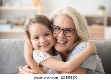 Happy old grandmother hugging little grandchild girl looking at camera, smiling mature mother or senior grandma granny laughing embracing adopted kid granddaughter sitting on couch, headshot portrait