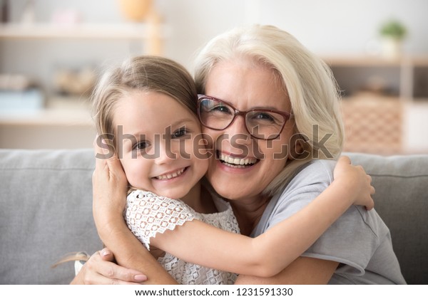 Happy old grandmother hugging little grandchild\
girl looking at camera, smiling mature mother or senior grandma\
granny laughing embracing adopted kid granddaughter sitting on\
couch, headshot portrait