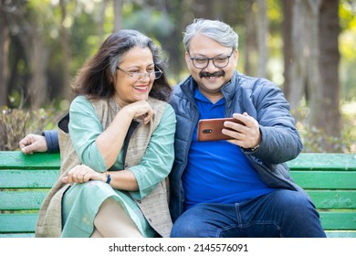 Happy Old  Indian Couple Using Smart Phone At Park, Old Man And Woman Watching Video Social Media On Mobile Phone While Sitting On Bench In The Garden.