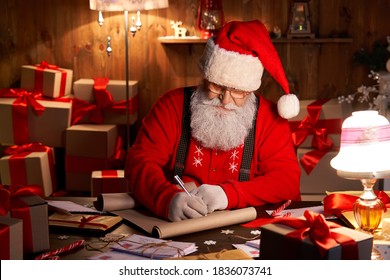Happy old kind bearded Santa Claus wearing hat, glasses, writing on wish list, working on Christmas eve sitting at cozy home workshop table late with presents, tree and candles preparing for holidays.