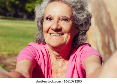 Happy Old Lady Taking Selfie While Walking In Park. Self Portrait Of Senior Grey Haired Woman In Casual With Grass And Trees In Background. Self Shot Concept