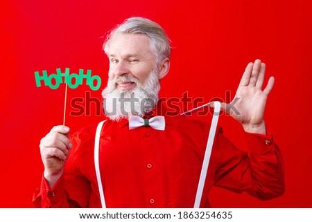 Happy, old man with long white beard and mustache posing, smiling, showing his suspenders  Positive modern male wearing stylish red shirt shows hohoho photo props. Winter holidays celebration concept.