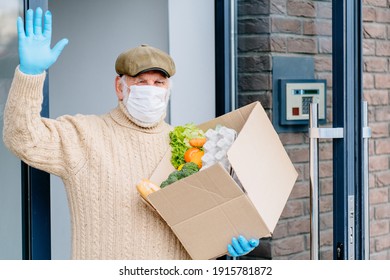 Happy Old Man In Medical Protective Mask And Gloves Holding Parcel With Grocery Fresh Food, Waving Hand In Doorway Of Her Apartment House, Outdoor. New Rules, Social Distance Helping Elderly People.
