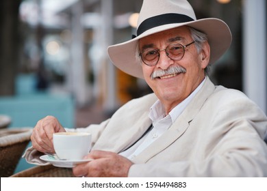 Happy Old Man On Vacation Drinking Coffee At Restaurant Enjoying Retirement Wearing White Suit And Hat