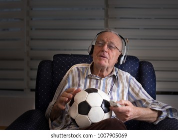 Happy Old Man With Remote Control And Soccer Ball Sitting In Armchair At Home And Watching Football Match