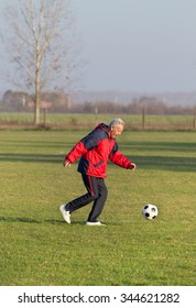 Happy Old Man In Seventies Kicking A Soccer Ball On The Grass Field