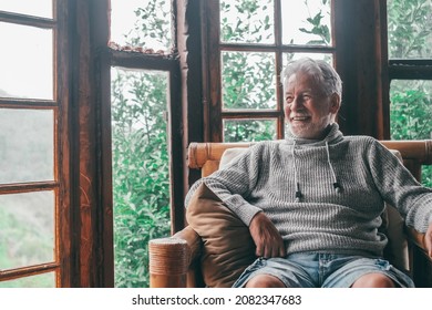 Happy Old Man Spending Leisure Time At Home Looking Out Through Window. Smiling Senior Man With Gray Hair And Sweater Relaxing On Wooden Armchair And Admiring View From Transparent Wooden Glass Door