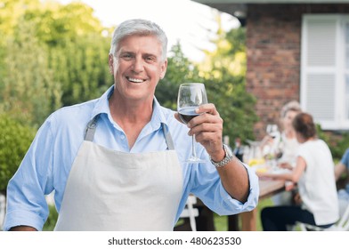 Happy Old Man Wearing Apron Raising A Toast. Senior Man Drinking A Glass Of Red Wine And Looking At Camera. Portrait Of Happy Grandfather Having Lunch With His Family Outside.