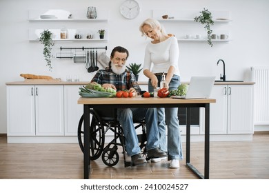 Happy old married man in wheelchair and wife prepare healthy salad, cutting thin slices of greenery vegetables and tomatoes with knife against background of spacious kitchen. - Powered by Shutterstock