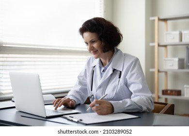Happy Old Mature 60s Female Doctor General Practitioner Doctor Physician Working On Computer, Handwriting Notes In Disease History On Clipboard Paper, Writing Healthcare Prescription In Clinic Office.