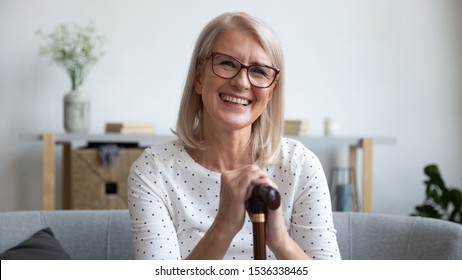 Happy old mature adult woman hold cane walking stick sit on sofa looking at camera, smiling senior retired middle aged grandmother satisfied with osteoporosis disease medical treatment, portrait
