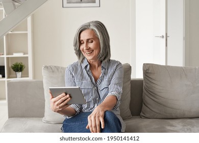 Happy old mature mid age senior woman using digital tablet looking at computer virtual calling having online meeting, watching tv, reading e book spending time with tech device sit on couch at home.
