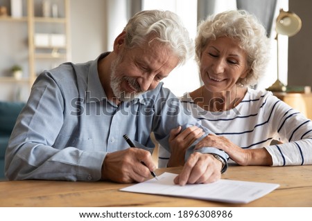 Happy old mature retired family couple putting signature on paper document, feeling satisfied with purchasing own apartment house or signing medical insurance agreement together at office meeting.