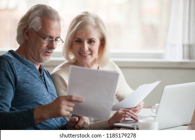 Happy old middle aged couple holding reading good news in document, smiling senior mature family excited by mail letter, checking paying domestic bills online on laptop, discussing budget planning