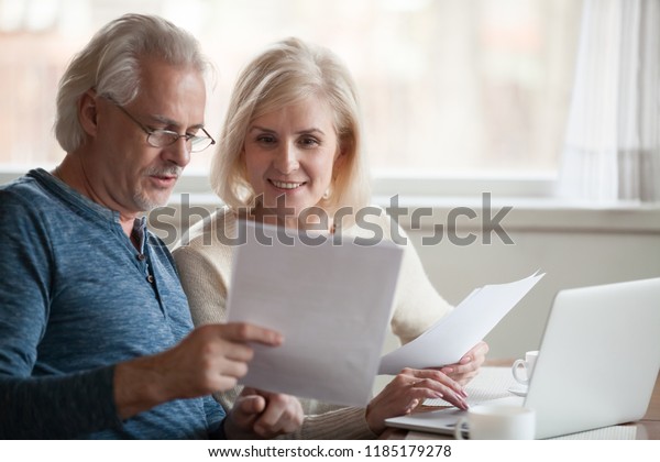 Happy old middle aged couple holding reading\
good news in document, smiling senior mature family excited by mail\
letter, checking paying domestic bills online on laptop, discussing\
budget planning