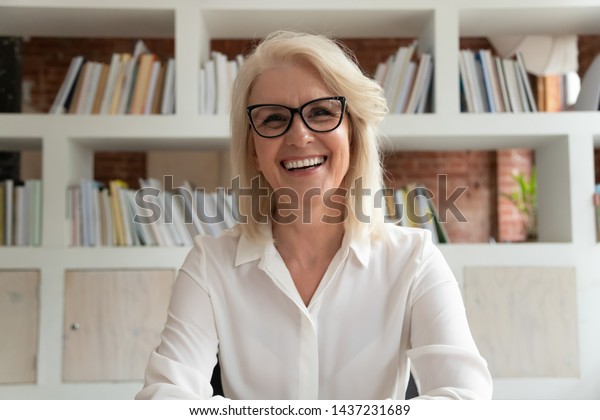 Happy old online teacher tutor look at camera\
give educational training class lecture teach by webcam, smiling\
mature business woman talk to webcam make video conference call sit\
at table in library