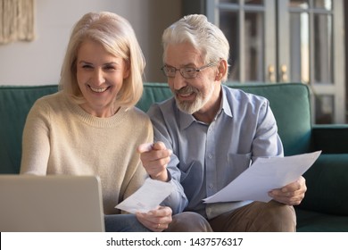 Happy Old Retired Couple Paying Domestic Bills Online Using Computer App, Satisfied Bank Clients Senior Middle Aged Grandparents Holding Papers Make Loan Insurance Payment At Home On Laptop On Sofa
