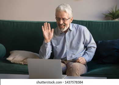 Happy Old Retired Man Waving Hand Talking To Webcam Make Distant Video Call Chat On Laptop Sit On Sofa, Cheerful Elder Grandpa Enjoy Online Communication Technology Looking At Computer Screen At Home