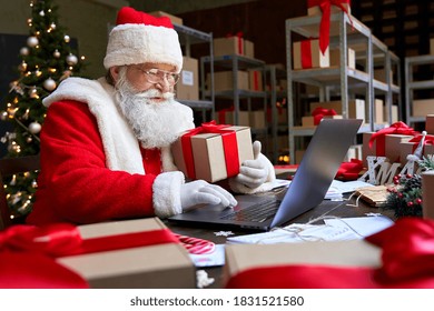 Happy old Santa Claus wearing costume holding gift box using laptop computer sitting at workshop table on Merry Christmas eve. Ecommerce website xmas time holiday online shopping sale concept. - Powered by Shutterstock