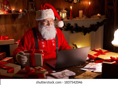 Happy old Santa Claus wearing hat holding gift box using laptop computer sitting at workshop home table late on Merry Christmas eve. Ecommerce website xmas time holiday online shopping e commerce sale