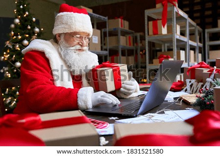 Happy old Santa Claus wearing costume holding gift box using laptop computer sitting at workshop table on Merry Christmas eve. Ecommerce website xmas time holiday online shopping sale concept.