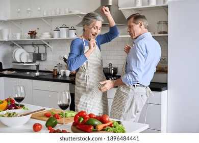 Happy old senior 50s couple having fun dancing cooking together in kitchen. Cheerful middle aged family wearing aprons drinking wine enjoying preparing healthy meal and celebrating anniversary at home