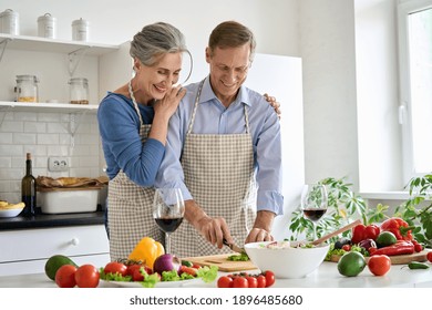 Happy Old Senior 50s Couple Wearing Aprons Having Fun Preparing Meal Salad At Home. Smiling Middle Aged Wife Hugging Older Husband Cutting Fresh Vegetables, Cooking Healthy Food Together In Kitchen.