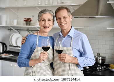 Happy Old Senior 50s Couple Wearing Aprons Hugging Having Fun Drinking Wine, Looking At Camera In Kitchen. Smiling Middle Aged Husband And Wife Holding Glasses, Cooking, Celebrating Together. Portrait