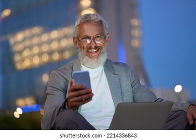 Happy Old Senior Business Man Professional, Older Businessman Sitting Outside Office In City Park Using Smartphone And Laptop Remote Working Online On Technology Devices Late At Night Outdoors.