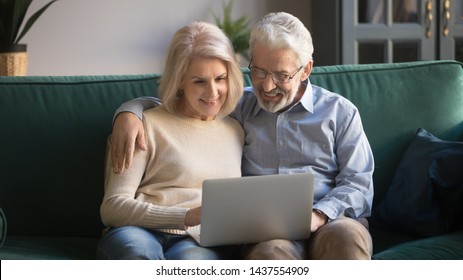 Happy Old Senior Couple Using Laptop Making Distance Video Call Chat Or Doing Internet Shopping Together, Retired Family Middle Aged Spouses Embracing Looking At Computer Screen Sit On Sofa At Home