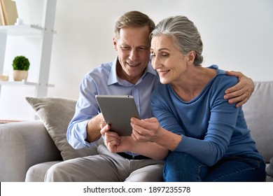 Happy old senior retired grandparents using digital tablet sitting on couch at home. Smiling mature 50s family couple enjoying websurfing online holding computer technology device making video call.