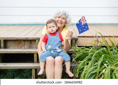 Happy Old Woman Grandmother With Grandson Baby Holding Australian Flag. Smiling Old Lady With Kid Child On Home Backyard Waving Australia Flag. Family Citizens Celebrating Australia Day.