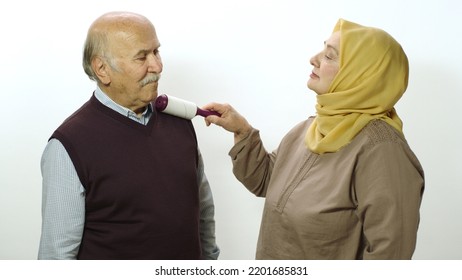 Happy Old Woman In Hijab Is Cleaning Pet Hair From Husband's Clothes With Sticky Clothing Roll.Studio Portrait Of Elderly Husband And Wife Isolated On White Background.Cat,dog And Pet Hair On Clothes.