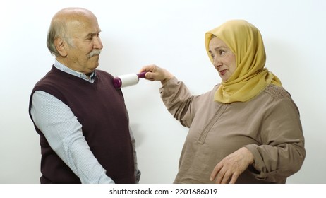 Happy Old Woman In Hijab Is Cleaning Pet Hair From Husband's Clothes With Sticky Clothing Roll.Studio Portrait Of Elderly Husband And Wife Isolated On White Background.Cat,dog And Pet Hair On Clothes.