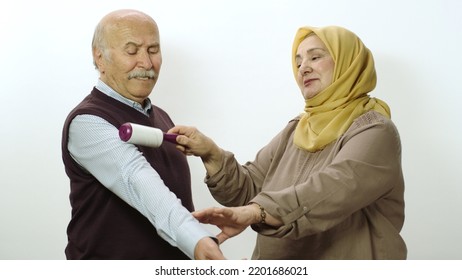 Happy Old Woman In Hijab Is Cleaning Pet Hair From Husband's Clothes With Sticky Clothing Roll.Studio Portrait Of Elderly Husband And Wife Isolated On White Background.Cat,dog And Pet Hair On Clothes.