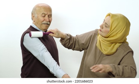 Happy Old Woman In Hijab Is Cleaning Pet Hair From Husband's Clothes With Sticky Clothing Roll.Studio Portrait Of Elderly Husband And Wife Isolated On White Background.Cat,dog And Pet Hair On Clothes.