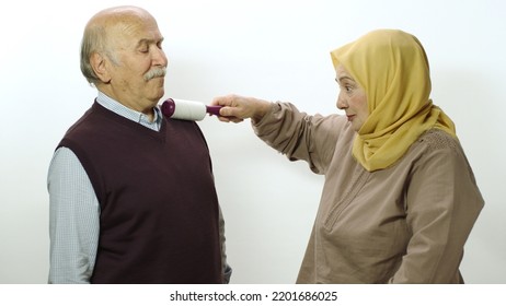 Happy Old Woman In Hijab Is Cleaning Pet Hair From Husband's Clothes With Sticky Clothing Roll.Studio Portrait Of Elderly Husband And Wife Isolated On White Background.Cat,dog And Pet Hair On Clothes.