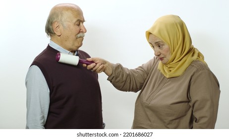 Happy Old Woman In Hijab Is Cleaning Pet Hair From Husband's Clothes With Sticky Clothing Roll.Studio Portrait Of Elderly Husband And Wife Isolated On White Background.Cat,dog And Pet Hair On Clothes.