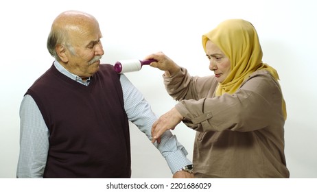 Happy Old Woman In Hijab Is Cleaning Pet Hair From Husband's Clothes With Sticky Clothing Roll.Studio Portrait Of Elderly Husband And Wife Isolated On White Background.Cat,dog And Pet Hair On Clothes.