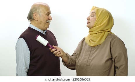 Happy Old Woman In Hijab Is Cleaning Pet Hair From Husband's Clothes With Sticky Clothing Roll.Studio Portrait Of Elderly Husband And Wife Isolated On White Background.Cat,dog And Pet Hair On Clothes.