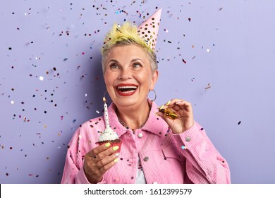 Happy Old Woman Looking At The Flying Confetti While Holding Cupcake, Isolated Over Violet Background