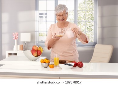 Happy old woman preparing healthy breakfast, pouring milk over cereals, smiling, looking at camera. - Powered by Shutterstock