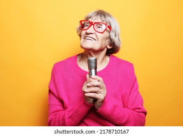 Happy Old Woman Singing With Microphone Over Yellow Color Background